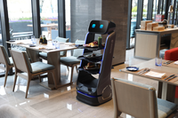 KBS Serviceroboter T5 in moderner Restaurantumgebung, befüllt mit Speisen auf drei Ebenen. Bildschirm mit blauem Gesichtsausdruck verleiht dem Roboter eine sympathische Wirkung.